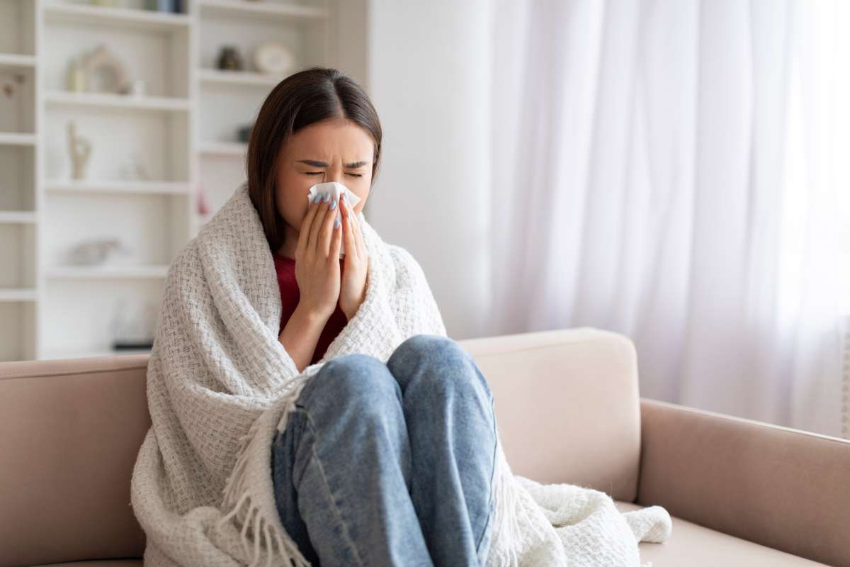 Young woman sits on the couch covered in a blanket while blowing her nose during flu season.